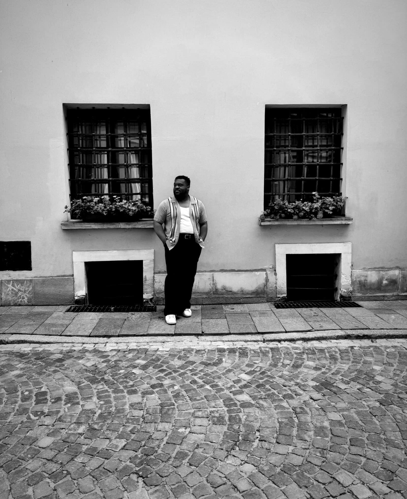 Zachary Stovall leaning against a wall between two barred windows on a cobblestone street, in black and white.